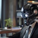 A humanoid robot wearing a business suit seated at an office desk with a laptop, a coffee cup, and a plant in the background.