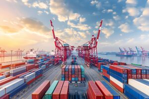 A bustling commercial shipping port with rows of colorful stacked containers, large red cranes, and container ships under a partly cloudy sky at sunset.