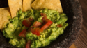 A close-up image of guacamole with tomato chunks served in a stone molcajete with tortilla chips arranged around the edges.