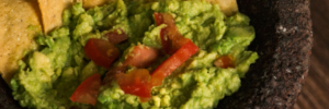 A close-up image of guacamole with tomato chunks served in a stone molcajete with tortilla chips arranged around the edges.