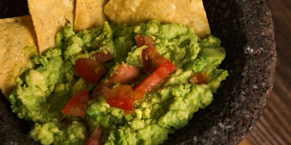 A close-up image of guacamole with tomato chunks served in a stone molcajete with tortilla chips arranged around the edges.