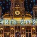 Illuminated signage "PARIS 2024" on a historically styled building facade with a clock, sculptures, and hanging flags at night.