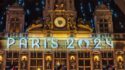 Illuminated signage "PARIS 2024" on a historically styled building facade with a clock, sculptures, and hanging flags at night.