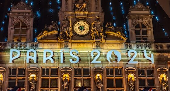 Illuminated signage "PARIS 2024" on a historically styled building facade with a clock, sculptures, and hanging flags at night.